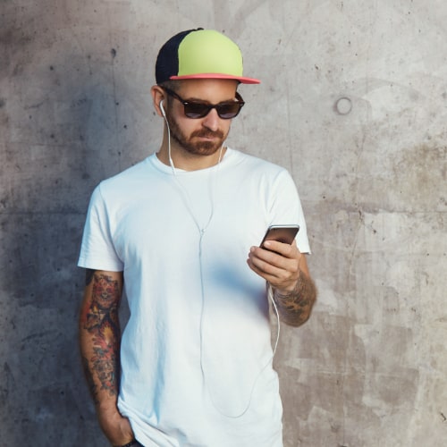 young-man-baseball-cap-sunglasses-white-blank-t-shirt-reading-something-his-smartphone-standing-gray-concrete-wall-min